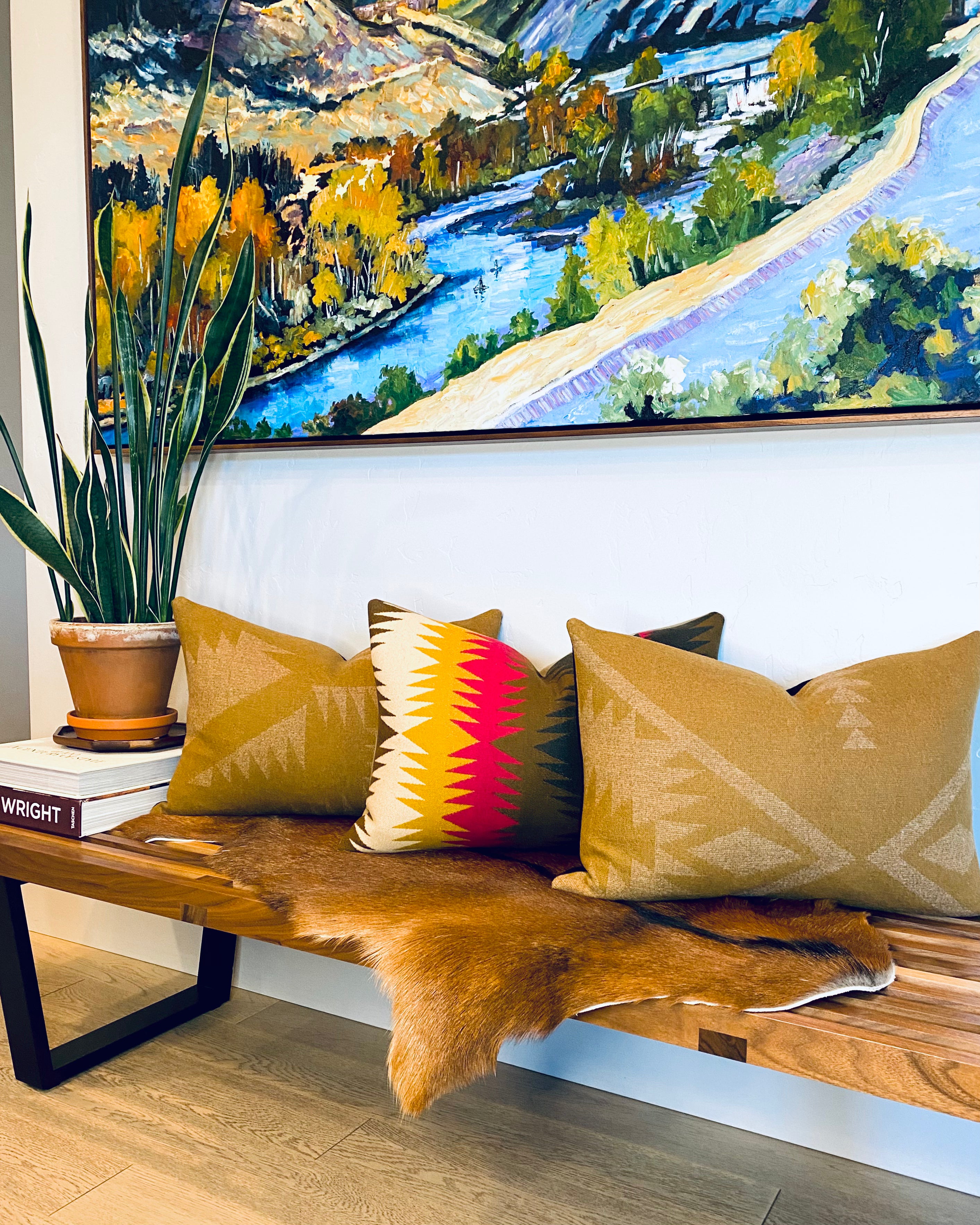A stylish entryway features a walnut George Nelson bench by Herman Miller, topped with a warm brown cowhide rug and three Pendleton wool accent pillows in golden-brown, red, olive, and yellow Southwestern designs. A snake plant and landscape painting complete this Pendleton home decor and designer accent pillow styled space.