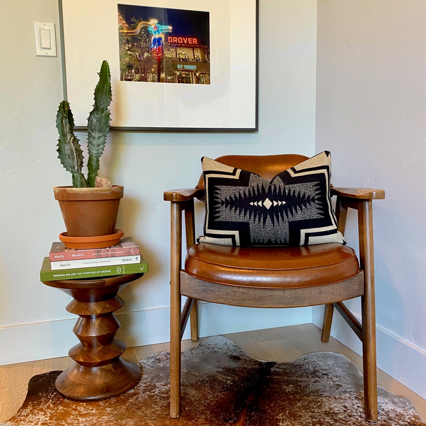 A cozy reading nook features a mid-century modern armchair, accented with a decorative throw pillow crafted from Pendleton wool fabric. The pillow showcases a prominent black and tan Southwestern geometric pattern. The chair sits on a brown and white cowhide rug. To the left, a potted cactus rests on a stack of design books, which are placed on an Eames walnut stool. Above, a framed photograph of a neon sign of "The Drover," hangs on a light gray wall. This image highlights Pendleton wool accent pillows.