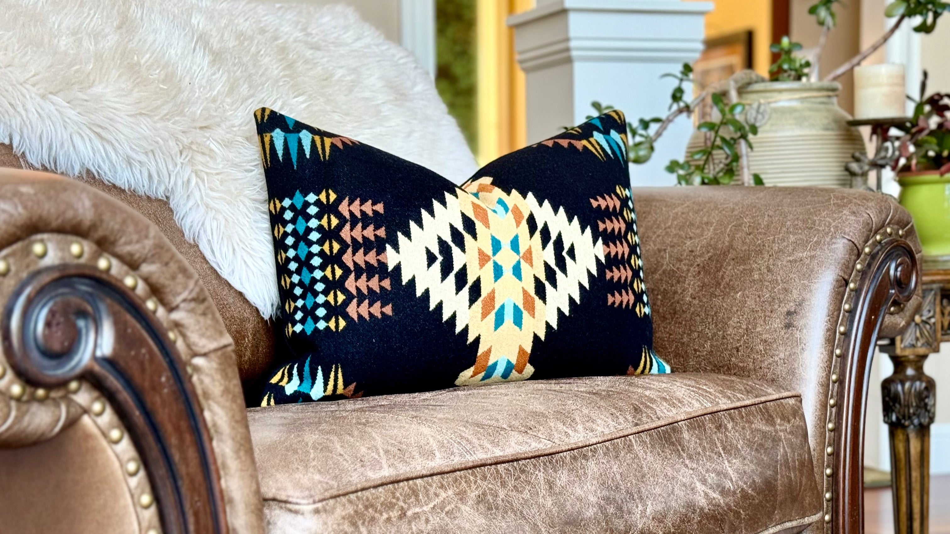 A rich, distressed brown leather armchair with nailhead trim and a white sheepskin throw features a vibrant Pendleton wool pillow. The black pillow showcases a striking Southwestern pattern in cream, orange, teal, and yellow. This inviting space highlights Pendleton wool home decor, Southwestern accent pillows, and luxury throw pillows for comfortable living in Boise, Idaho.