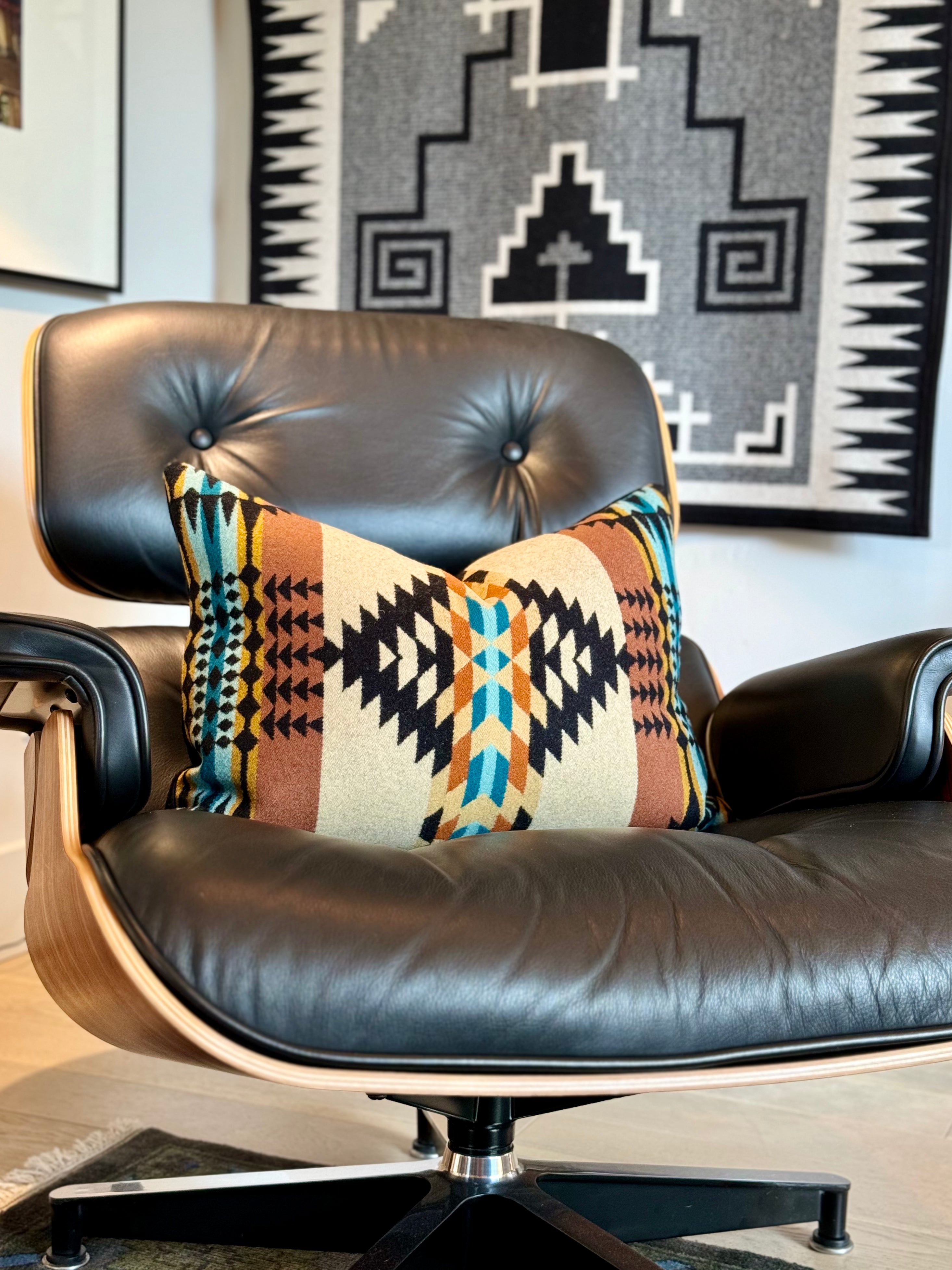 An Eames lounge chair, upholstered in black leather with a walnut wood molded frame, holds a decorative throw pillow crafted from Pendleton wool fabric. The pillow features a Southwestern-inspired design with a cream-colored center band, bordered by patterns in shades of brown, black, teal, and orange. In the background, a black and white geometric pattern Pendleton saddle blanket hangs on the wall. This image showcases Pendleton wool home decor and luxury accent pillows.