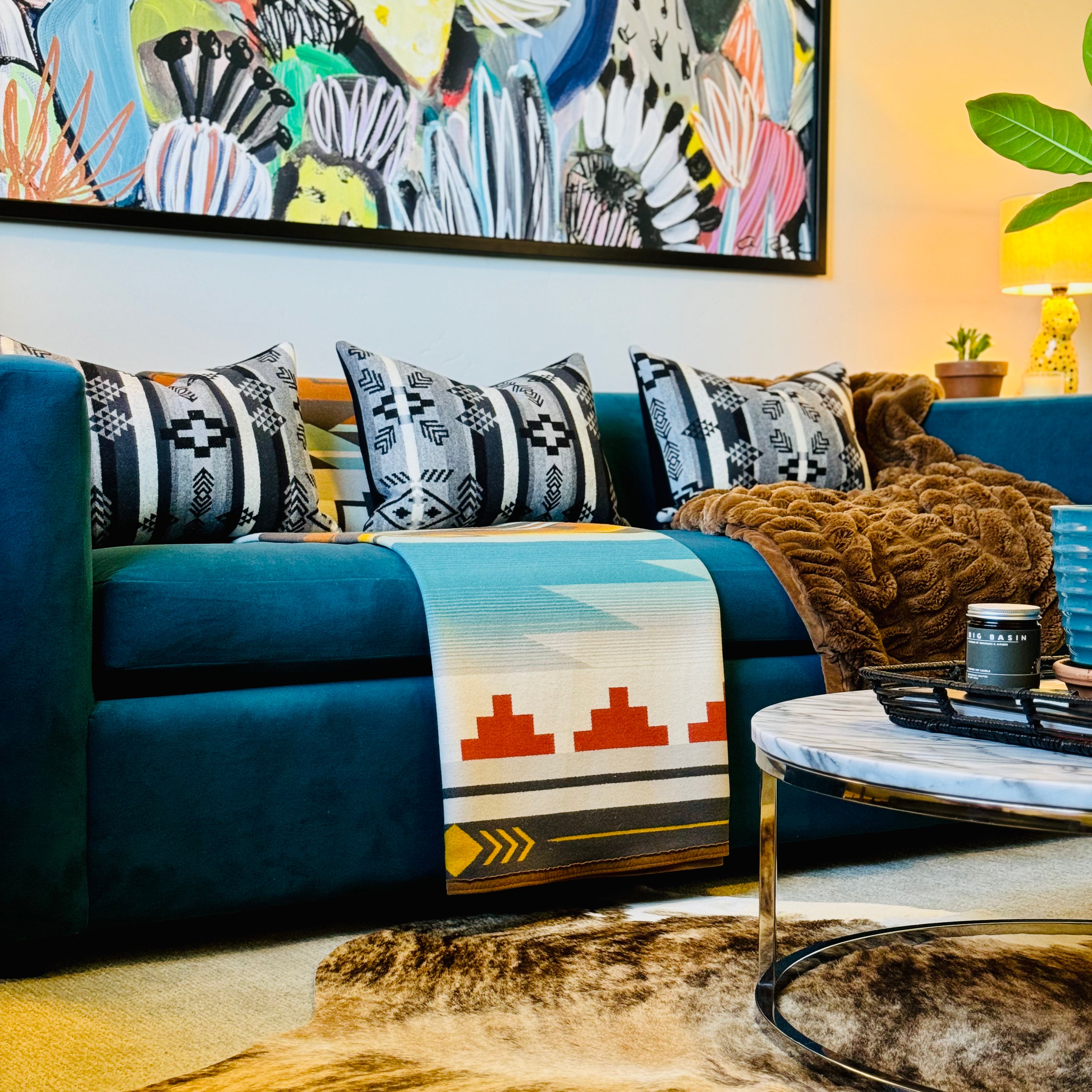A modern living room features a vibrant teal sofa, adorned with three decorative throw pillows crafted from Pendleton wool fabric in black, white, and gray Southwestern geometric patterns. A colorful Pendleton wool blanket with a prominent Southwestern design in blues, oranges, and white is draped over the sofa, next to a cozy brown textured throw. A light brown cowhide rug lies on the floor in front of the sofa. Above the sofa hangs a large, colorful abstract painting with floral or cactus-like motifs.