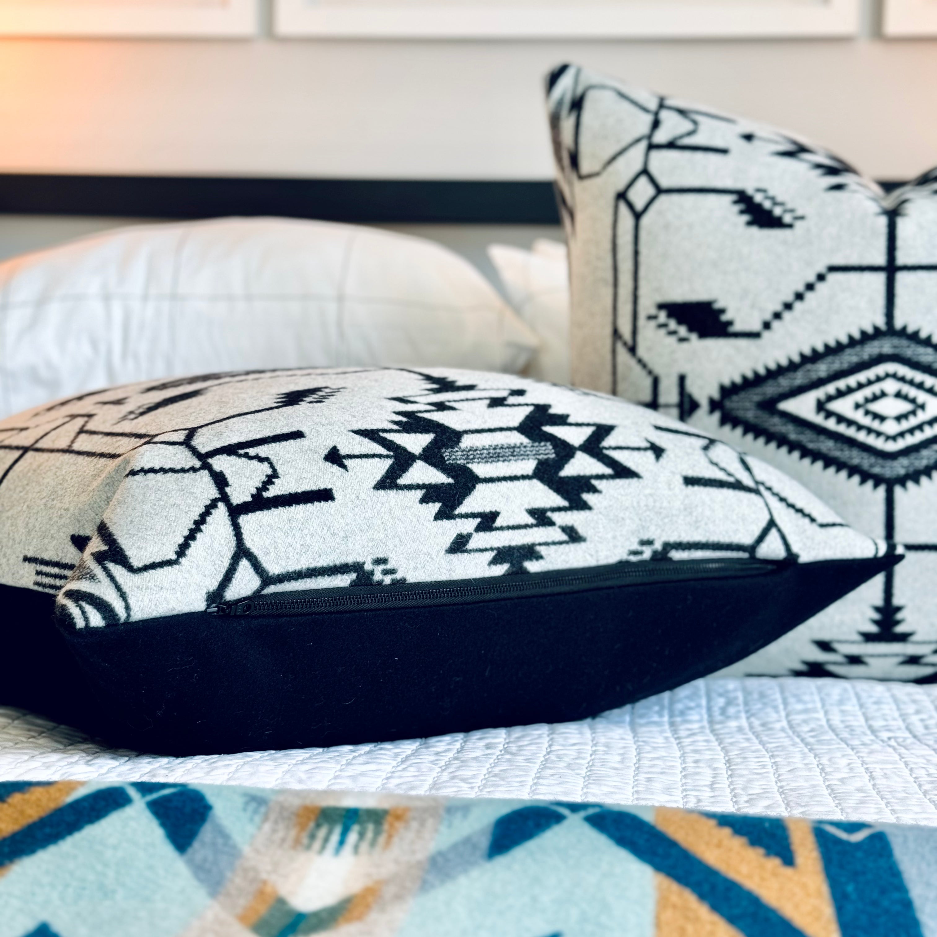 A neatly made bed features two decorative throw pillows crafted from Pendleton wool fabric, displaying a prominent black and white Southwestern geometric pattern. The foreground shows a partial view of a colorful Pendleton wool blanket or throw in shades of blue, yellow, and white, also with a Southwestern design. The bed is adorned with crisp white linens, and behind it, framed artwork with text "BOISE IDAHO," "McCALL IDAHO" is visible. This image highlights Pendleton wool accent pillows and blankets.