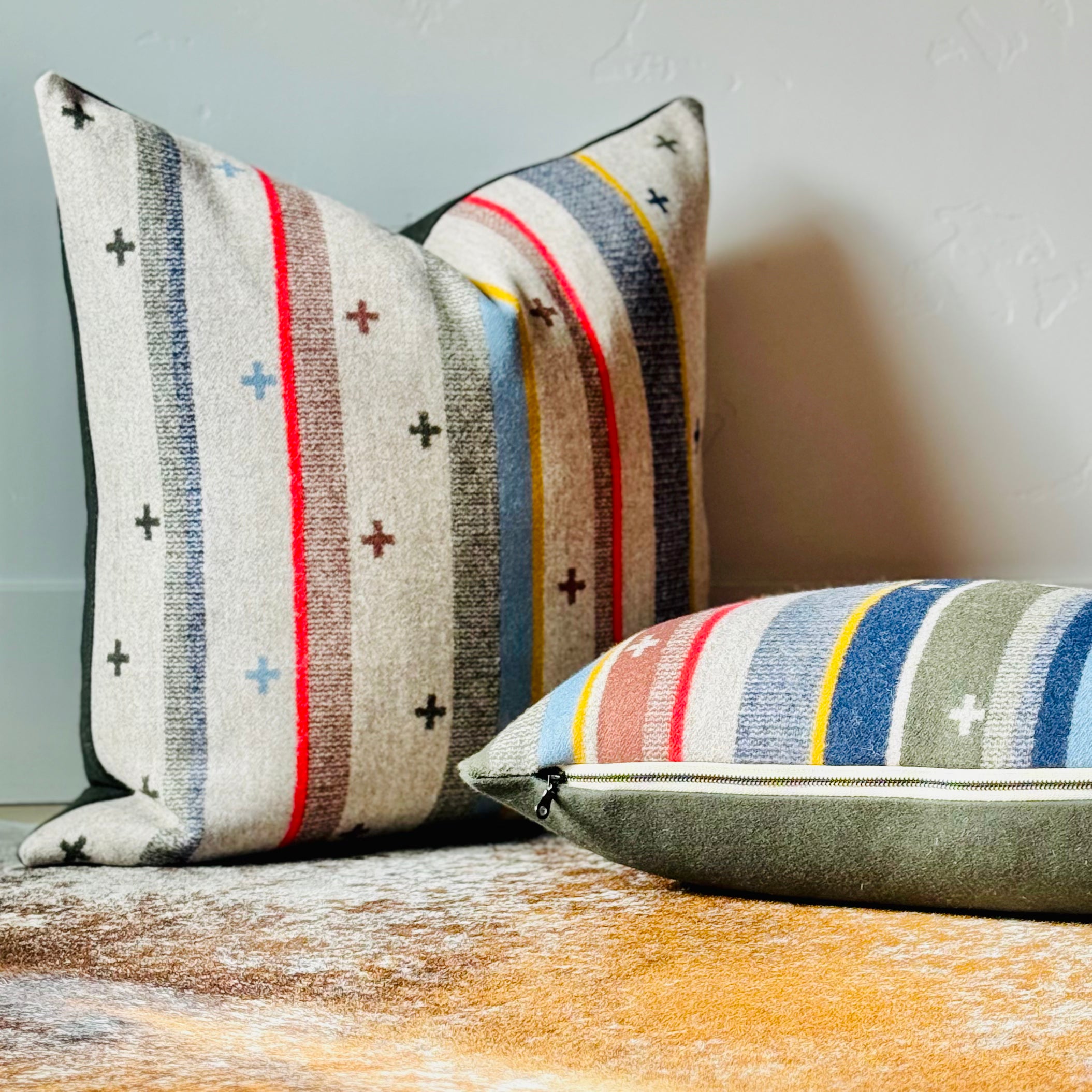 Two decorative throw pillows, crafted from Pendleton wool fabric, are displayed on a warm brown and white cowhide rug. The pillows feature a light taupe base with vertical stripes in various colors, including red, yellow, blue, and olive interspersed with small "plus" symbols. The back of the pillows appears to be a solid olive fabric. This image highlights Pendleton wool throw pillows, Southwestern home decor, and designer accent pillows for sale in Boise, Idaho.